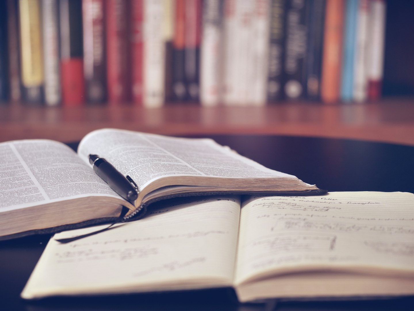 An open book and notebook with a pen on a dark wooden desk.